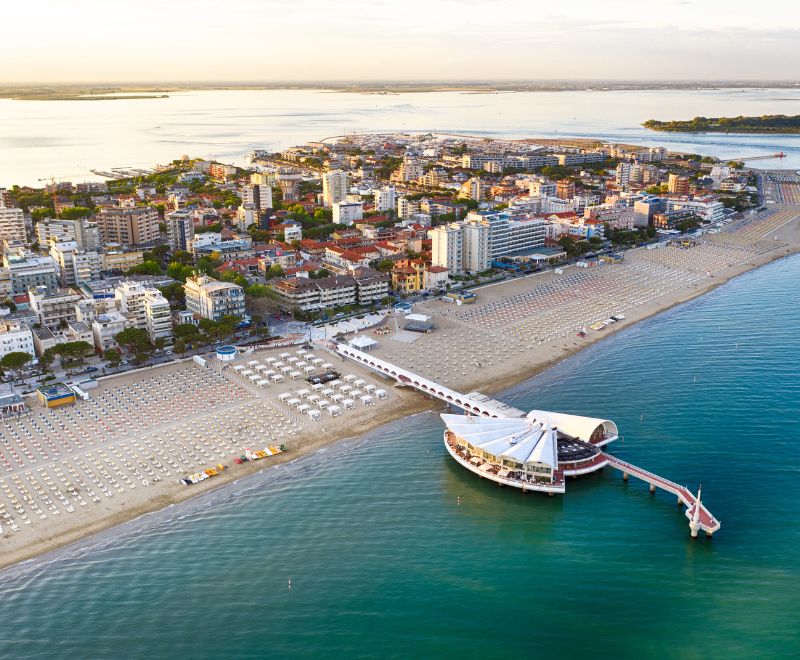 Lignano Sabbiadoro vista dall’alto: la lunga spiaggia dorata, ordinata con i suoi ombrelloni, incontra il mare Adriatico. In primo piano la celebre Terrazza a Mare, simbolo della località balneare, tra vivacità estiva e paesaggi unici.