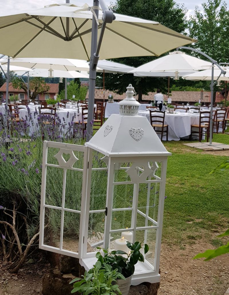 Un angolo suggestivo del giardino dell’Osteria Solder: una lanterna bianca decorata con cuori si apre tra lavanda e verde, mentre sullo sfondo tavoli imbanditi sotto grandi ombrelloni attendono gli ospiti per un’esperienza conviviale e raffinata.